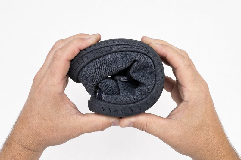 Ein flacher Minimalschuh aus dunkelgrauem Textil wird von zwei Händen so zusammengerollt, dass die Zehenkappe die Ferse berührt und ein geschlossener Ring entsteht; die dünne biegsame Gummisohle verläuft sichtbar an der Außenseite des Rings